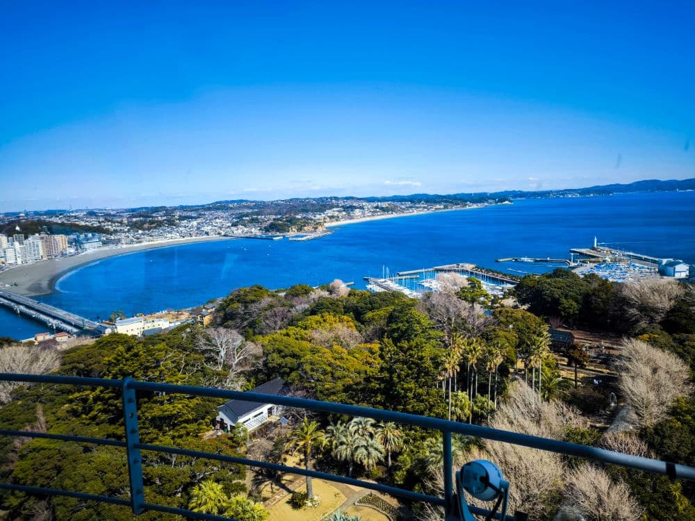 Ein Panoramablick auf eine Küstenstadt mit einer geschwungenen Bucht, einer langen Brücke, Jachthäfen, grünen Wäldern und Gebäuden unter einem strahlend blauen Himmel, aus dem sich die ikonische Enoshima-Seekerze erhebt.
