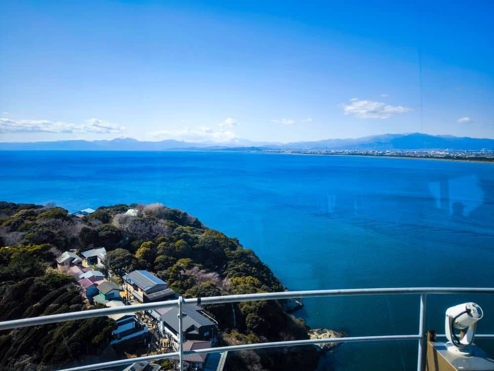 Eine malerische Aussicht von einem hohen Aussichtspunkt in der Nähe der Enoshima-Meereskerze zeigt den tiefblauen Ozean, die grünen Gebäude auf einer Halbinsel und die entfernten Berge unter einem klaren blauen Himmel, mit einem Geländer und einem münzbetriebenen Zuschauer im Vordergrund.