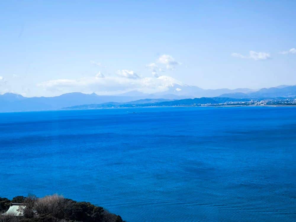 Ein leuchtend blauer Ozean erstreckt sich in Richtung einer entfernten Küste, mit sanften Hügeln und Bergen im Hintergrund unter einem klaren Himmel. Der schneebedeckte Berg Fuji und die ikonische Enoshima-Seekerze sind in der Nähe des Horizonts zu sehen.