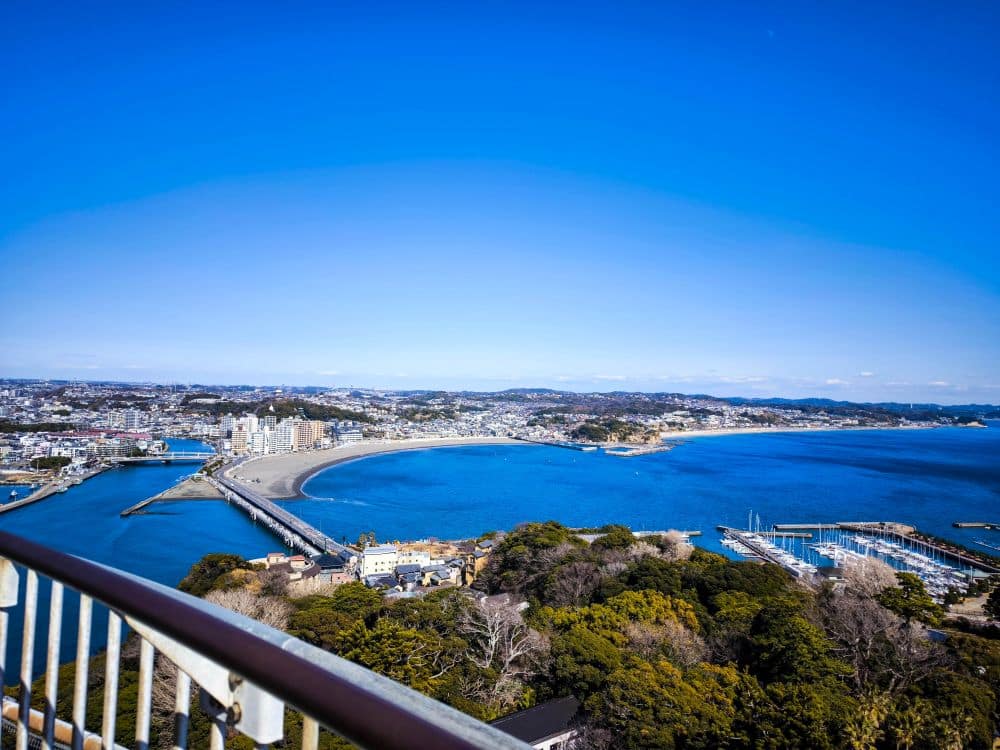 Der Blick von der Enoshima-Meereskerze zeigt eine Küstenstadt mit einem geschwungenen Sandstrand, blauem Meerwasser, einem mit Booten besetzten Jachthafen und üppig grünen Bäumen im Vordergrund unter einem klaren blauen Himmel.