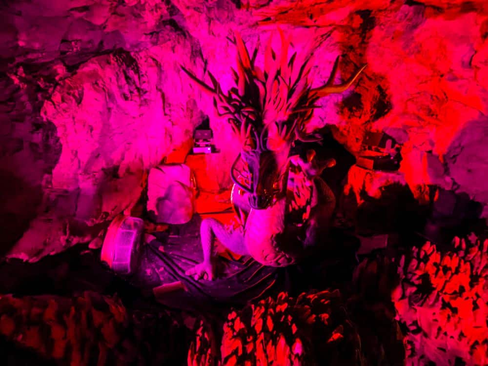 Eine Drachenstatue mit großen Hörnern sitzt in einer felsigen Iwaya-Höhle, die von roten und rosafarbenen Lichtern dramatisch beleuchtet wird und eine intensive und mystische Atmosphäre schafft.