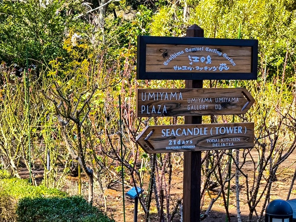 Ein hölzerner Wegweiser in einem Garten weist auf den Umiyama-Platz, die Umiyama-Galerie und die Enoshima-Meereskerze hin; üppige grüne Büsche und Bäume bilden den Hintergrund.