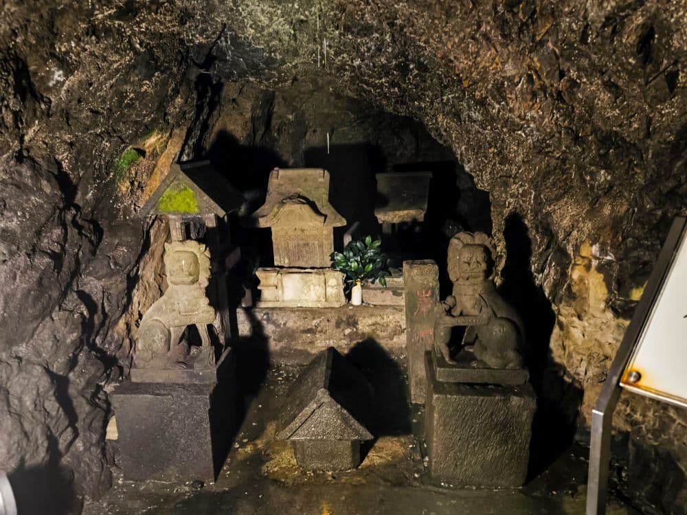 In der schwach beleuchteten Iwaya-Höhle stehen Steinstatuen und kleine Altäre, und zwischen den Felsen sind Moos und Pflanzen zu sehen, die eine geheimnisvolle und heilige Atmosphäre schaffen.