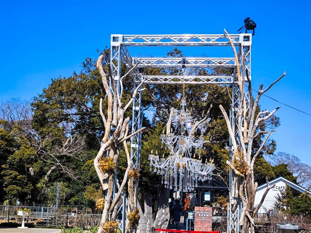 Ein großer Kristallkronleuchter hängt im Freien an einem mit Treibholz verzierten Metallrahmen vor grünen Bäumen und blauem Himmel in der Nähe der Enoshima-Meereskerze. Auf dem Gestell sitzt ein schwarzer Vogel, und rechts davon steht ein kleines Gebäude.