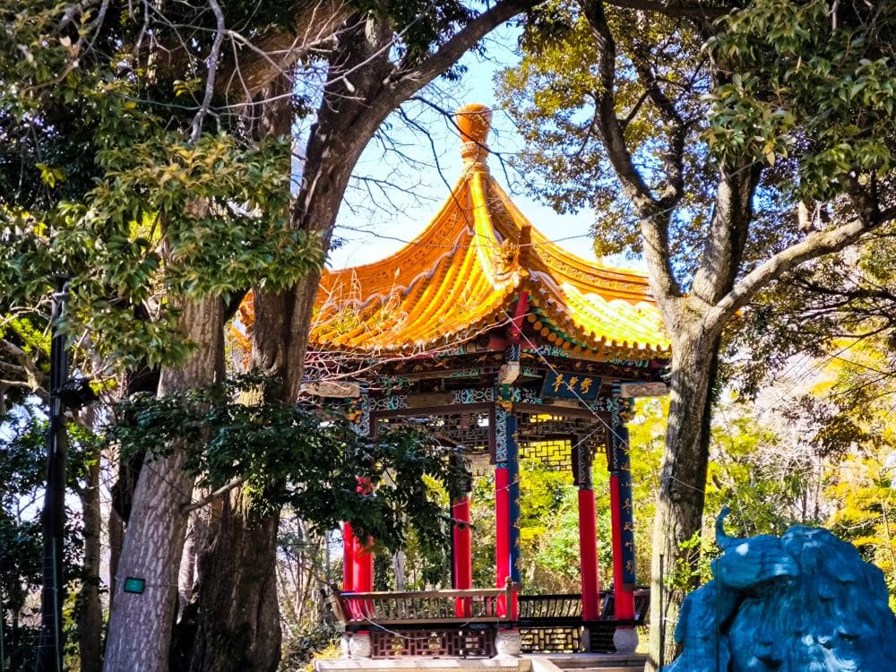 Ein traditioneller chinesischer Pavillon mit einem goldenen, verschnörkelten Dach und roten Säulen steht inmitten hoher, grüner Bäume im hellen Sonnenlicht, mit der Enoshima-Meereskerze im fernen Hintergrund.