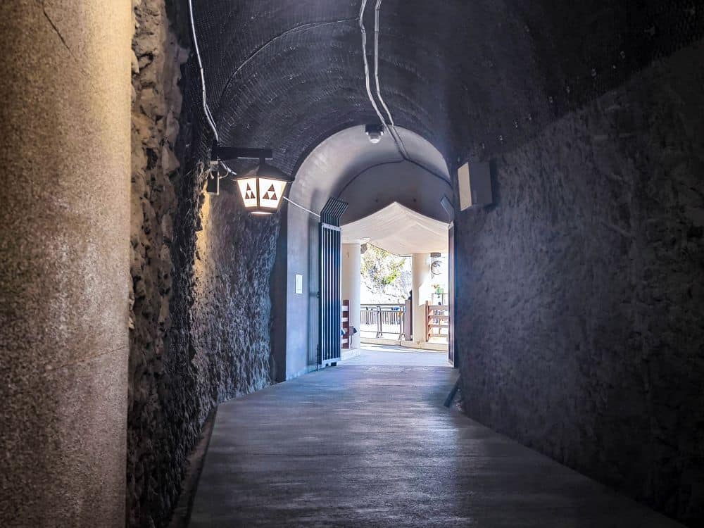 Ein schummrig beleuchteter Tunnel mit Steinwänden führt zu einem hellen Außenbereich. Zwei Laternen hängen an der linken Wand, und das Sonnenlicht am Ausgang von Iwaya gibt den Blick frei auf eine Terrasse mit Tischen und Stühlen, die in warmes Licht getaucht sind.