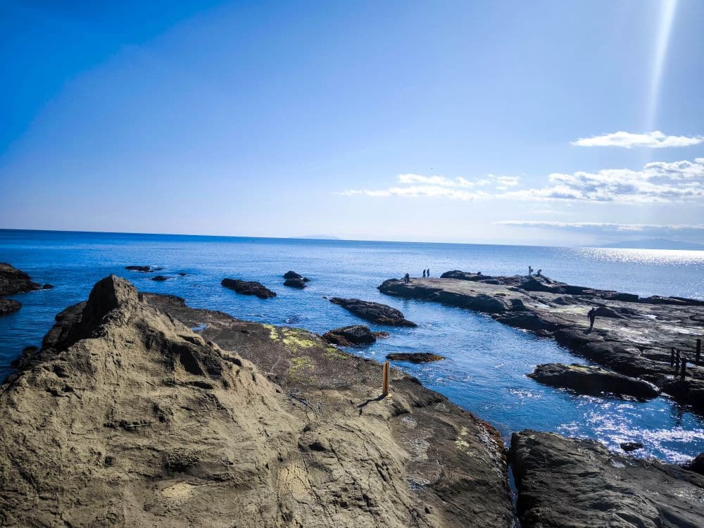 Felsige Küste mit großen Iwaya-Steinformationen im Vordergrund und Menschen, die auf den Felsen am blauen Meer stehen, unter einem hellen, klaren Himmel, in dem sich das Sonnenlicht auf dem Wasser spiegelt.