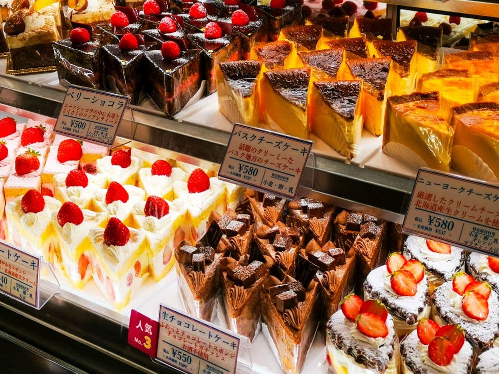 Eine Vitrine mit verschiedenen Kuchen, darunter Kurisumasukēki und japanische Weihnachtskuchen mit Erdbeeren und Schokolade, alle mit Preisschildern und Etiketten auf Japanisch.