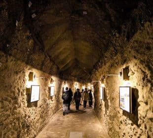 Eine Gruppe von Menschen geht durch den schwach beleuchteten Iwaya-Steintunnel mit seinen gewölbten Decken und gerahmten Bildern an den Wänden.