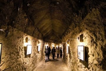 Eine Gruppe von Menschen geht durch den schwach beleuchteten Iwaya-Steintunnel mit seinen gewölbten Decken und gerahmten Bildern an den Wänden.