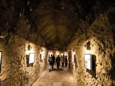 Eine Gruppe von Menschen geht durch den schwach beleuchteten Iwaya-Steintunnel mit seinen gewölbten Decken und gerahmten Bildern an den Wänden.