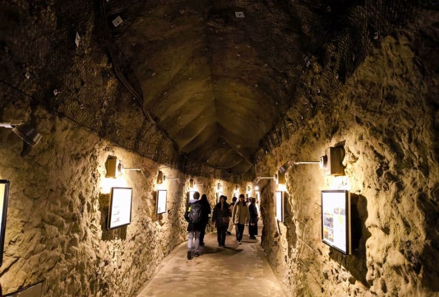 Eine Gruppe von Menschen geht durch den schwach beleuchteten Iwaya-Steintunnel mit seinen gewölbten Decken und gerahmten Bildern an den Wänden.
