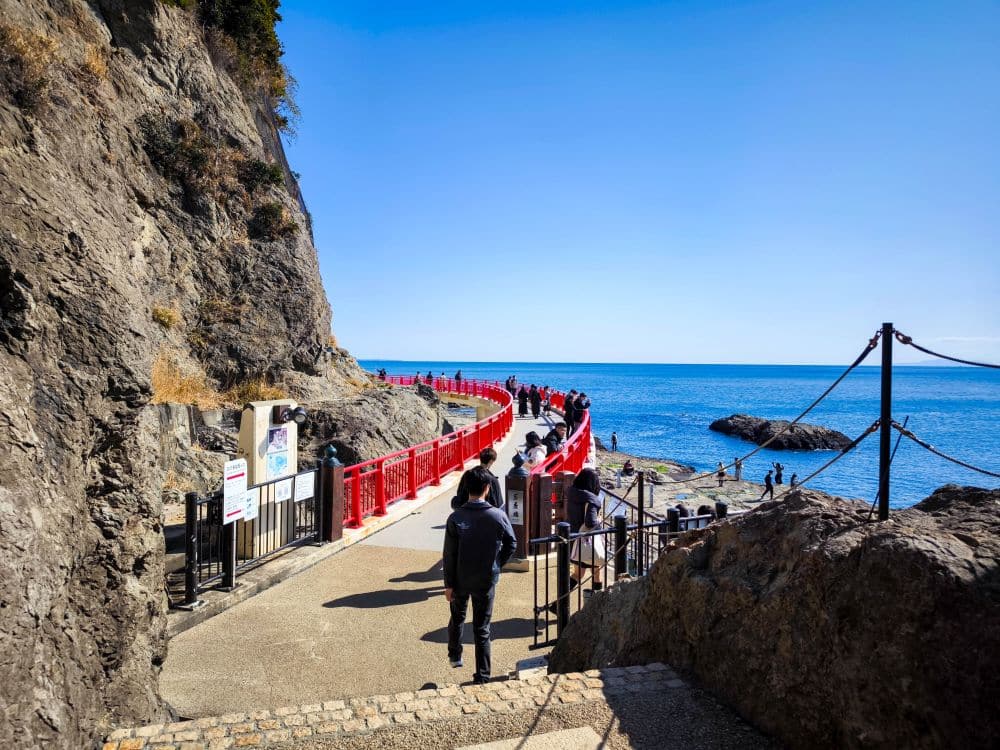 Auf dem geschwungenen Iwaya-Pfad mit leuchtend rotem Geländer spaziert man an felsigen Klippen und dem blauen Meer unter einem klaren Himmel entlang. Einige Besucher befinden sich in der Nähe des Wassers, und am Eingang zu dieser landschaftlich reizvollen Route sind Schilder angebracht.