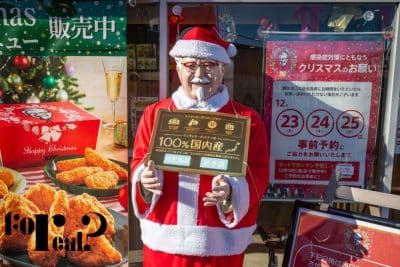 Eine lebensgroße Colonel-Sanders-Statue als Weihnachtsmann verkleidet steht vor einem KFC in Japan und hält ein Schild in Japanisch. Weihnachtsdekoration und Werbung für Brathähnchengerichte unterstreichen die Tradition des japanischen Weihnachtsessens bei KFC.