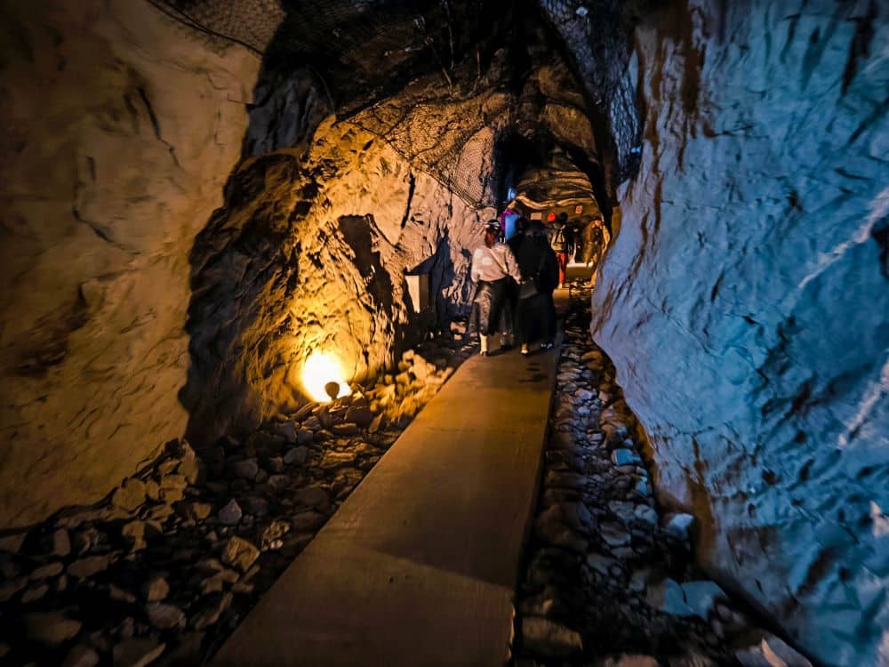 Die Menschen gehen durch die schwach beleuchtete Iwaya, einen felsigen unterirdischen Tunnel mit unebenen Steinwänden und einem schmalen Weg; ein Licht erhellt einen Teil des Ganges.
