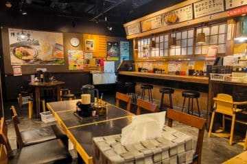 Ein gemütliches japanisches Ramen-Restaurant mit Holztischen und -stühlen, einer Taschentuchbox auf dem Tisch, einem Tresen mit Hockern und warmer Beleuchtung - perfekt für ein authentisches Restaurantbesuch-Japan-Erlebnis.