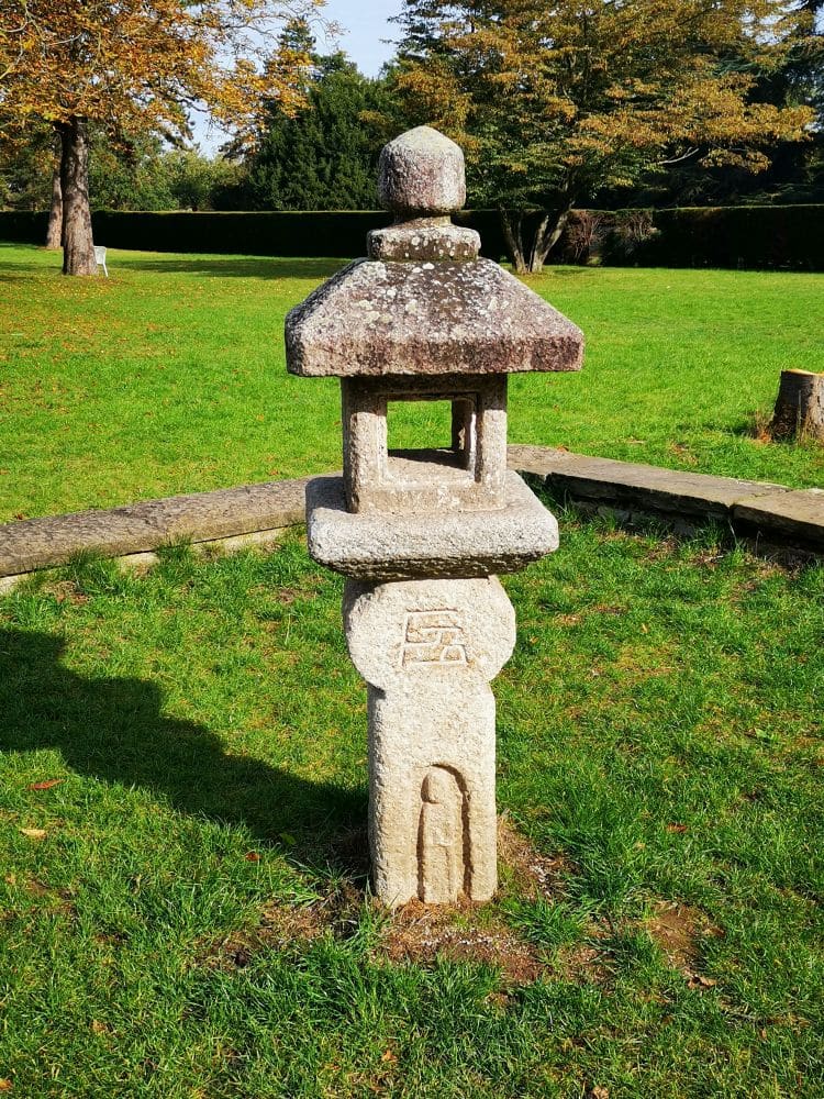 Eine traditionelle japanische Steinlaterne steht auf einer Wiese in einem sonnigen Park, umgeben von Bäumen und einer geschwungenen Steinbank. Sie erinnert an die japanische Atmosphäre des Japanischen Gartens im Nordpark Düsseldorf.