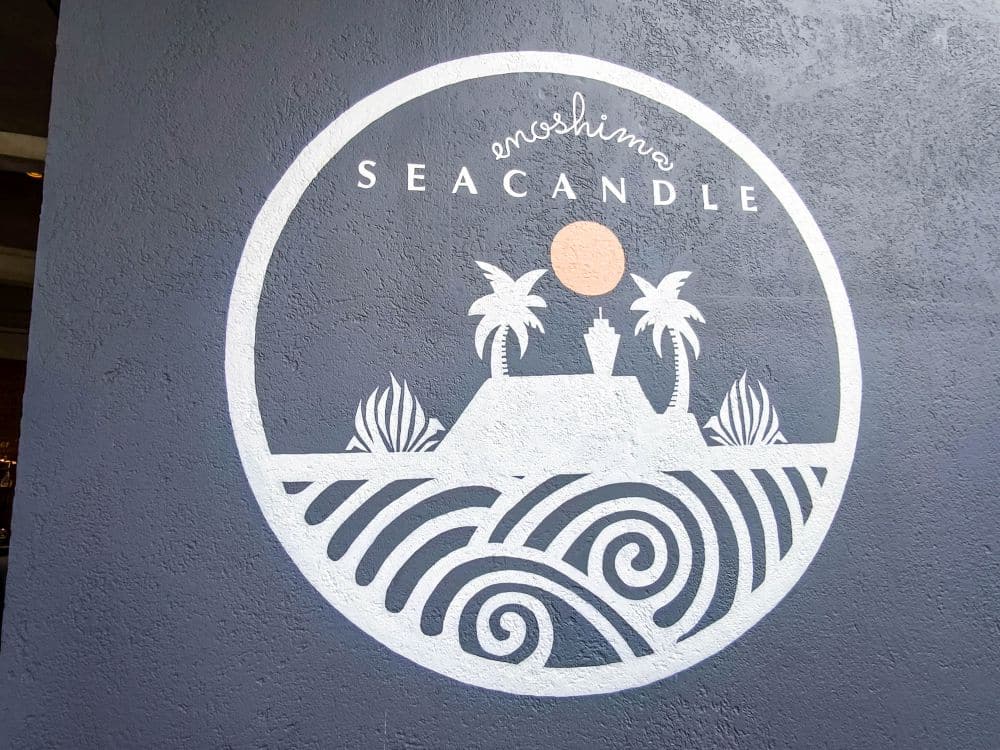 Ein weißes, kreisförmiges Wandbild auf einer grauen Wand zeigt den Text "Enoshima Sea Candle" und eine Insel mit einem Leuchtturm, Palmen, Wellen und einer stilisierten Sonne.