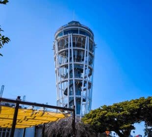 Ein hoher, weißer Aussichtsturm mit einem offenen Metallgerüst und einer Wendeltreppe - bekannt als die Enoshima-Meereskerze - steht vor einem klaren blauen Himmel, umgeben von Bäumen und einer gelben Überdachung im Vordergrund.