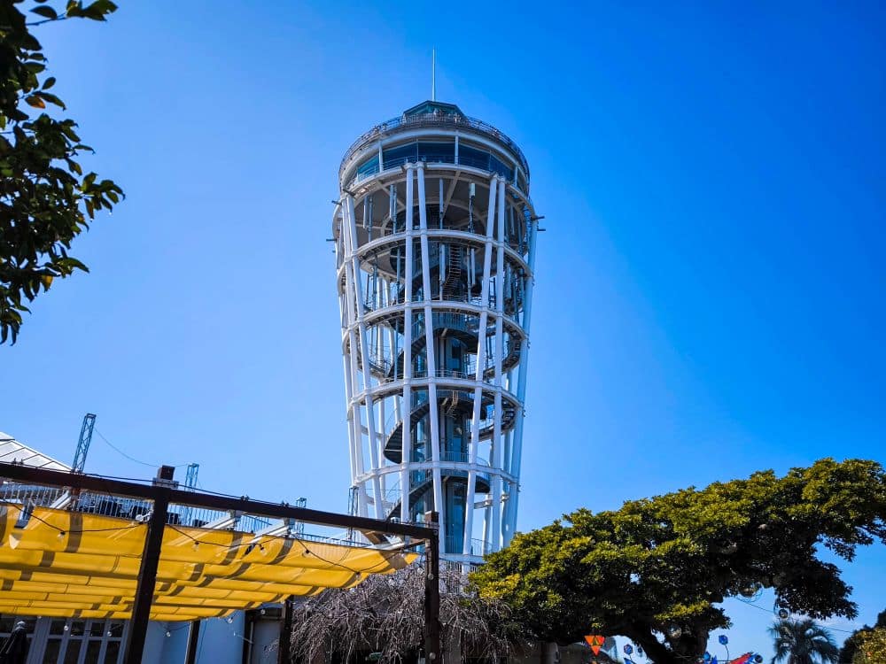Ein hoher, weißer Aussichtsturm mit einem offenen Metallgerüst und einer Wendeltreppe - bekannt als die Enoshima-Meereskerze - steht vor einem klaren blauen Himmel, umgeben von Bäumen und einer gelben Überdachung im Vordergrund.