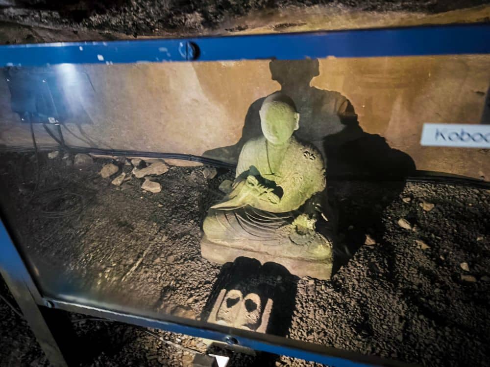 Eine steinerne Statue einer sitzenden Figur in Gewändern, ausgestellt hinter Glas in einer schwach beleuchteten, felsigen Iwaya-Umgebung. Die Statue wirft einen Schatten an die Wand, und vor ihr sind zwei steinerne Fußabdrücke zu sehen.