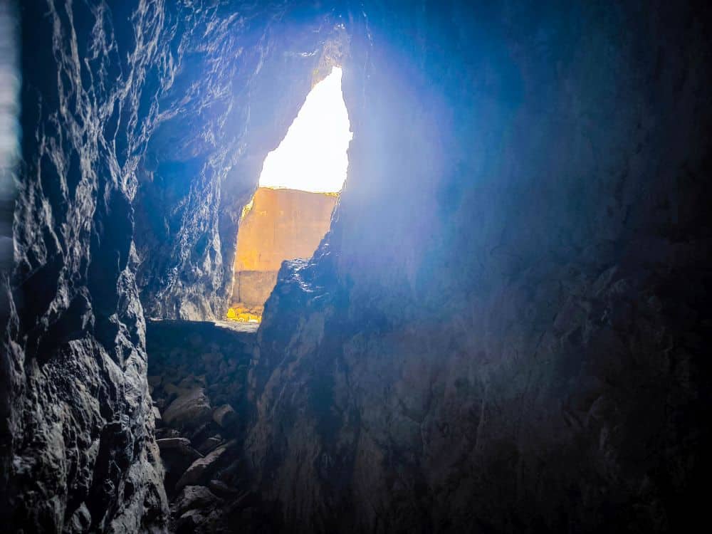 Blick aus dem Inneren der dunklen, felsigen Iwaya-Höhle auf eine helle, dreieckige Öffnung, in die Sonnenlicht einfällt und einen Teil der zerklüfteten Wände und des Bodens in der Nähe des Eingangs beleuchtet.