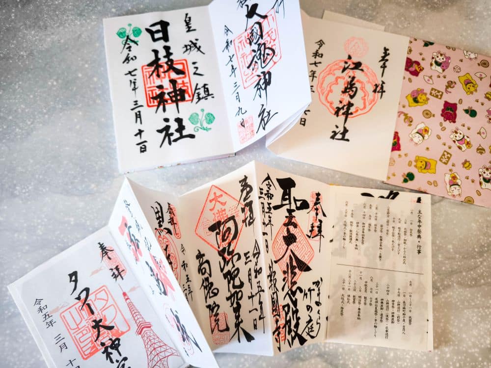 Mehrere aufgeschlagene japanische Goshuincho (Tempel-Stempelbücher) mit komplizierter Kalligrafie, roten Goshuin-Stempeln und bunten Einbänden sind auf einer hellen, gesprenkelten Oberfläche ausgestellt. Ein Buch enthält Aufkleber mit niedlichen Figuren.