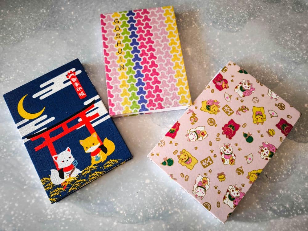Drei bunte Notizbücher sind auf einer gesprenkelten Oberfläche ausgestellt. Eines ähnelt einem Goshuin-Buch mit Füchsen und einem Torii-Tor, ein anderes hat ein geometrisches Regenbogenmuster, und das dritte ist mit Glückskatzen und Rosen verziert.