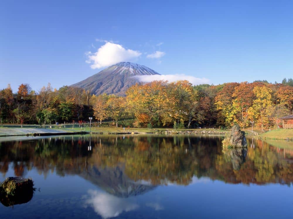 In einem ruhigen See spiegeln sich leuchtendes Herbstlaub und farbenfrohe Herbstbäume wider, mit einem schneebedeckten Berg unter einem klaren blauen Himmel und ein paar Wolken, die um den Gipfel des Berges treiben.