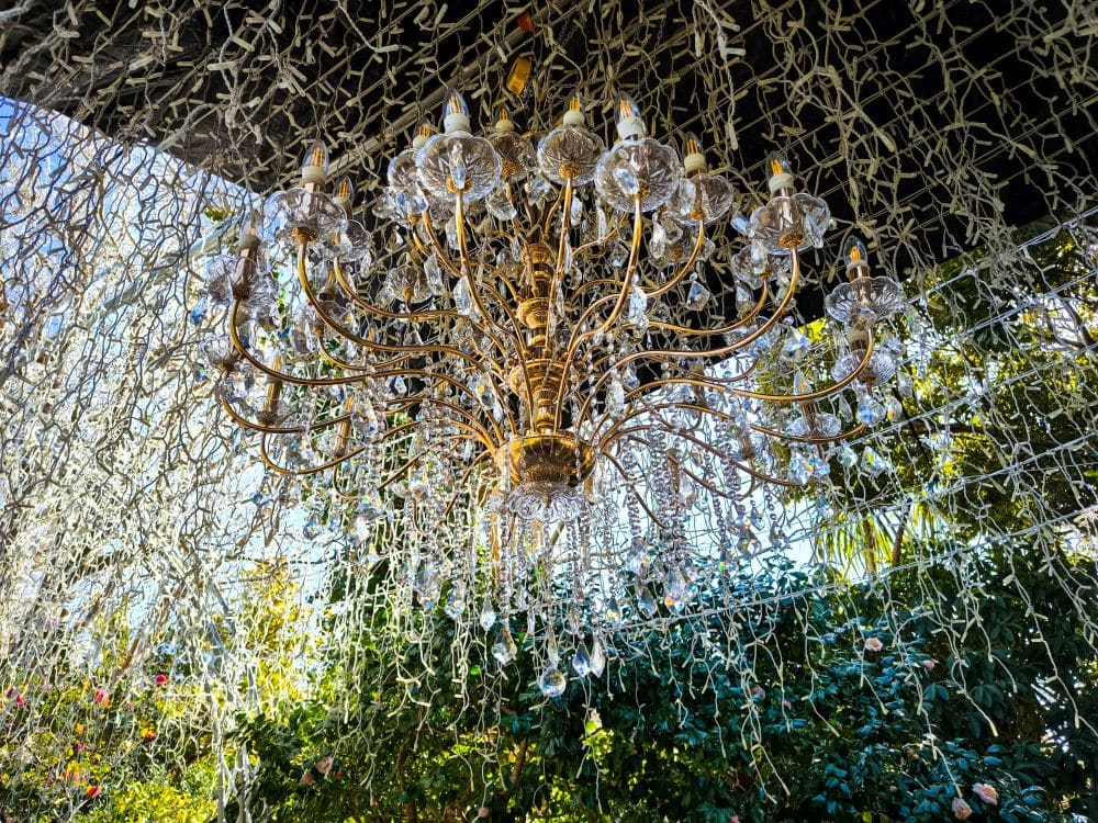 Ein goldener Kronleuchter mit Glasakzenten hängt im Freien unter einem Baldachin aus Lichterketten, während sich im Hintergrund Grünpflanzen und die Enoshima-Meereskerze gegen den blauen Himmel abheben.