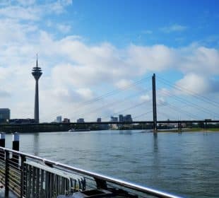 Blick auf den Rhein in Düsseldorf, Deutschland, mit dem Rheinturm, der Oberkasseler Brücke, modernen Gebäuden und einem teilweise bewölkten blauen Himmel - perfekt, um Düsseldorfs japanisches Flair und eine kulinarische Reise entlang des Ufers zu genießen.