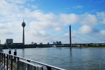 Blick auf den Rhein in Düsseldorf, Deutschland, mit dem Rheinturm, der Oberkasseler Brücke, modernen Gebäuden und einem teilweise bewölkten blauen Himmel - perfekt, um Düsseldorfs japanisches Flair und eine kulinarische Reise entlang des Ufers zu genießen.