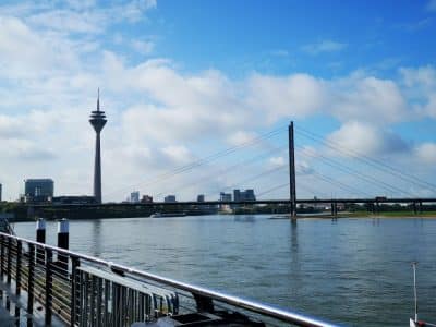 Blick auf den Rhein in Düsseldorf, Deutschland, mit dem Rheinturm, der Oberkasseler Brücke, modernen Gebäuden und einem teilweise bewölkten blauen Himmel - perfekt, um Düsseldorfs japanisches Flair und eine kulinarische Reise entlang des Ufers zu genießen.