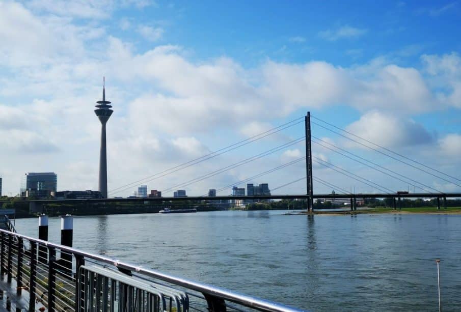 Blick auf den Rhein in Düsseldorf, Deutschland, mit dem Rheinturm, der Oberkasseler Brücke, modernen Gebäuden und einem teilweise bewölkten blauen Himmel - perfekt, um Düsseldorfs japanisches Flair und eine kulinarische Reise entlang des Ufers zu genießen.