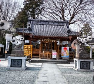 Ein traditioneller japanischer Kumano-Schrein mit einem Ziegeldach und einer Holzstruktur, flankiert von Steinstatuen und weißen Laternen. Bäume umgeben den Schrein, und in der Nähe hängen Ziertafeln und Glückspapiere.