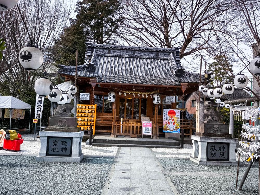 Ein traditioneller japanischer Kumano-Schrein mit einem Ziegeldach und einer Holzstruktur, flankiert von Steinstatuen und weißen Laternen. Bäume umgeben den Schrein, und in der Nähe hängen Ziertafeln und Glückspapiere.