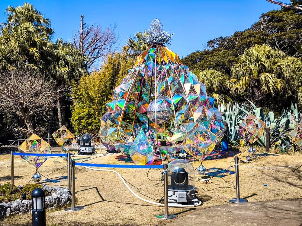 Eine lebendige, geometrische Kunstinstallation aus schillernden Paneelen steht auf einer Wiese in der Nähe der Enoshima-Meereskerze, umgeben von tropischen Pflanzen und Bäumen unter einem strahlend blauen Himmel, mit Lichtern und Geräten drum herum.