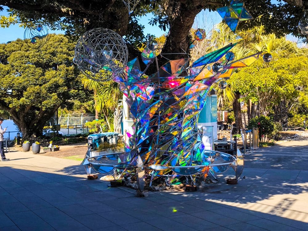 Die farbenfrohe, reflektierende geometrische Skulptur aus Glas oder Kunststoff steht im Freien in der Nähe der Enoshima-Meereskerze, umgeben von Bäumen, Pflanzen und einem gepflasterten Gehweg. Das Sonnenlicht erzeugt Regenbogenreflexe auf der Skulptur und dem Boden.