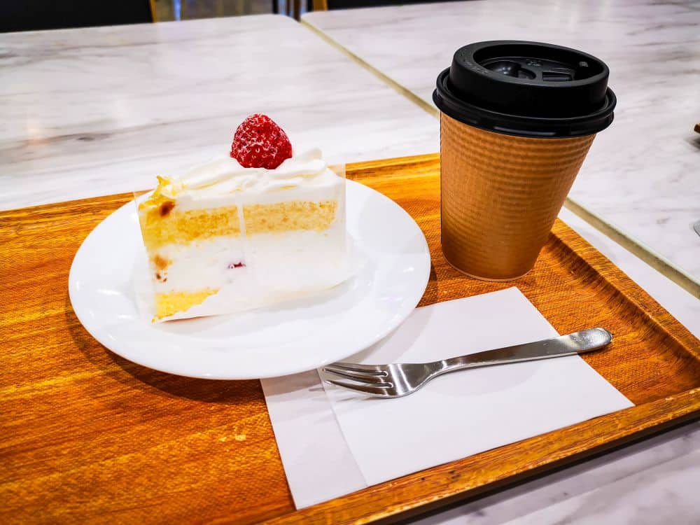 Ein Stück Kurisumasukēki, die klassische japanische Weihnachtstorte, mit Sahne und einer Erdbeere obendrauf, liegt auf einem weißen Teller neben einer Gabel, daneben eine Kaffeetasse zum Mitnehmen auf einem Holztablett.