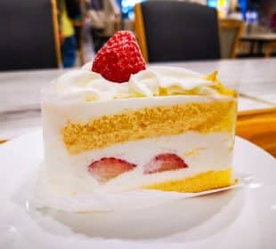 Ein Stück Kurisumasukēki, der kultige japanische Weihnachtskuchen, mit Biskuit und Schlagsahne, gekrönt von einer ganzen Erdbeere, liegt auf einem weißen Teller in einem hell erleuchteten Café.