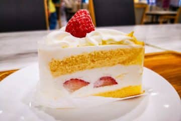 Ein Stück Kurisumasukēki, der kultige japanische Weihnachtskuchen, mit Biskuit und Schlagsahne, gekrönt von einer ganzen Erdbeere, liegt auf einem weißen Teller in einem hell erleuchteten Café.