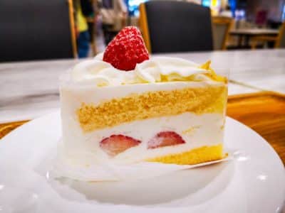 Ein Stück Kurisumasukēki, der kultige japanische Weihnachtskuchen, mit Biskuit und Schlagsahne, gekrönt von einer ganzen Erdbeere, liegt auf einem weißen Teller in einem hell erleuchteten Café.