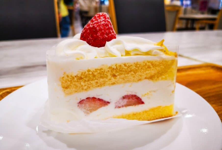 Ein Stück Kurisumasukēki, der kultige japanische Weihnachtskuchen, mit Biskuit und Schlagsahne, gekrönt von einer ganzen Erdbeere, liegt auf einem weißen Teller in einem hell erleuchteten Café.