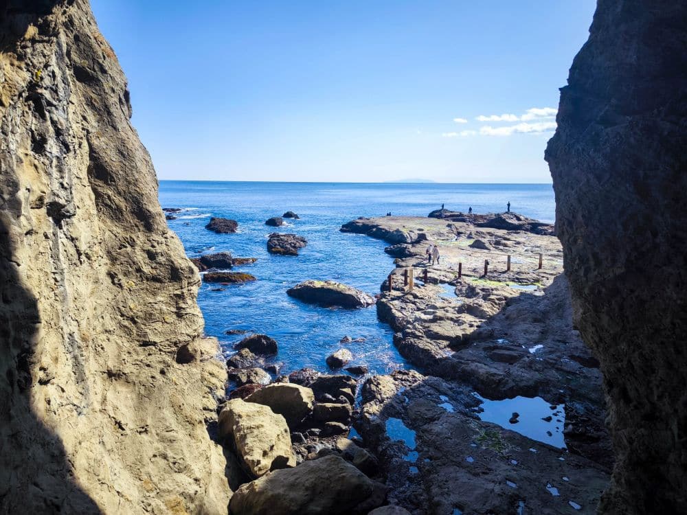 Blick aus dem Inneren der Iwaya-Höhle auf den strahlend blauen Ozean mit verstreuten Felsen und Gezeitentümpeln am Ufer. Einige Menschen stehen auf dem Felsvorsprung unter einem klaren Himmel.