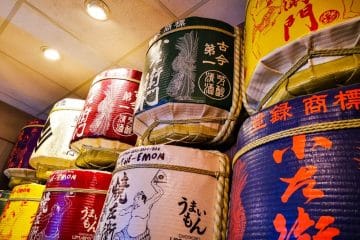 In einem Innenraum in Little Tokyo stehen stapelweise bunte, große, traditionelle japanische Sakefässer mit kunstvollen Kalligraphien und Illustrationen, die dicht an dicht an einer Decke mit warmer Beleuchtung angeordnet sind.