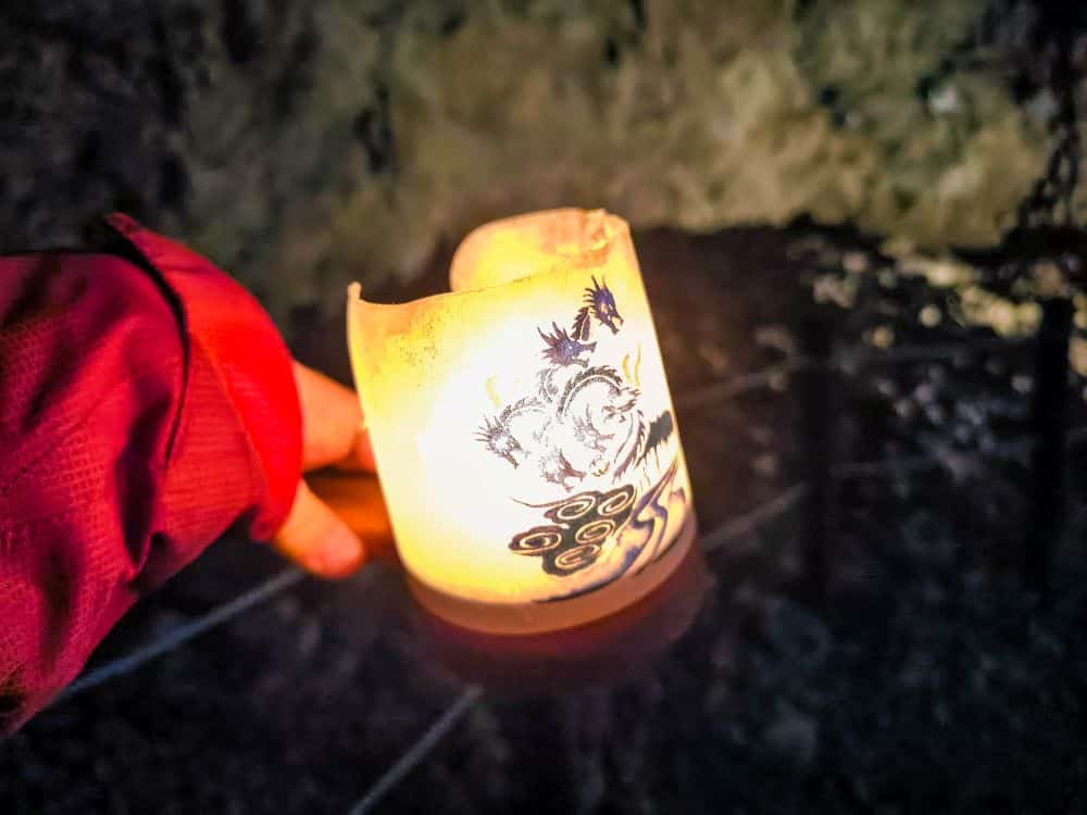 Eine Hand in einem roten Ärmel hält eine leuchtende Kerze mit dekorativen Mustern, die den dunklen, felsigen Iwaya-Hintergrund beleuchtet.