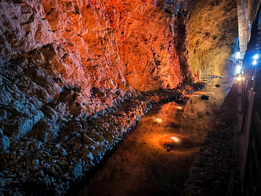 Der Iwaya-Tunnel zeichnet sich durch felsige, orange beleuchtete Wände und einen flachen, mit Wasser gefüllten Boden aus, in dem sich die Beleuchtung spiegelt. Kleine Lichter erhellen den Gang, während am anderen Ende helles Licht winkt.