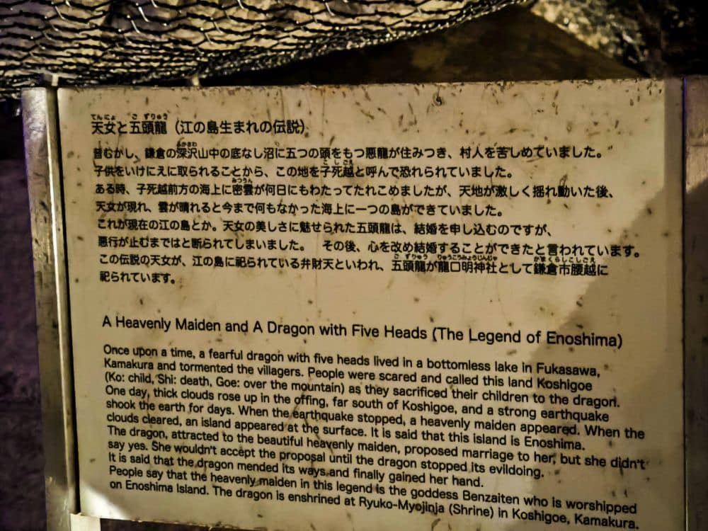 Ein verwittertes Schild in der Nähe der Iwaya-Höhle mit Text in Japanisch und Englisch erzählt die Legende von Enoshima, in der ein Mädchen und ein fünfköpfiger Drache beschrieben werden. Flecken und Markierungen verdecken Teile des englischen Textes, so dass er nur teilweise lesbar ist.
