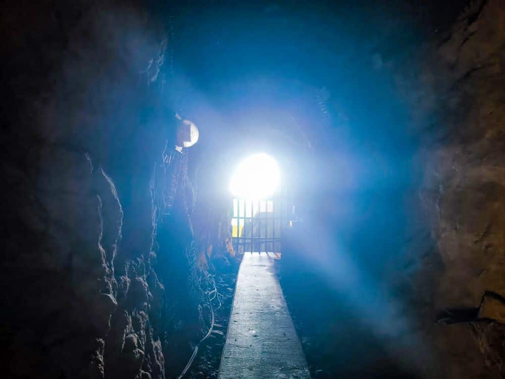 Blick aus dem Inneren von Iwaya, einem dunklen, felsigen Tunnel, auf ein helles Licht am Eingang, wo ein Metalltor den Weg nach draußen versperrt. Blaues Licht strömt herein und erhellt teilweise die rauen Wände.