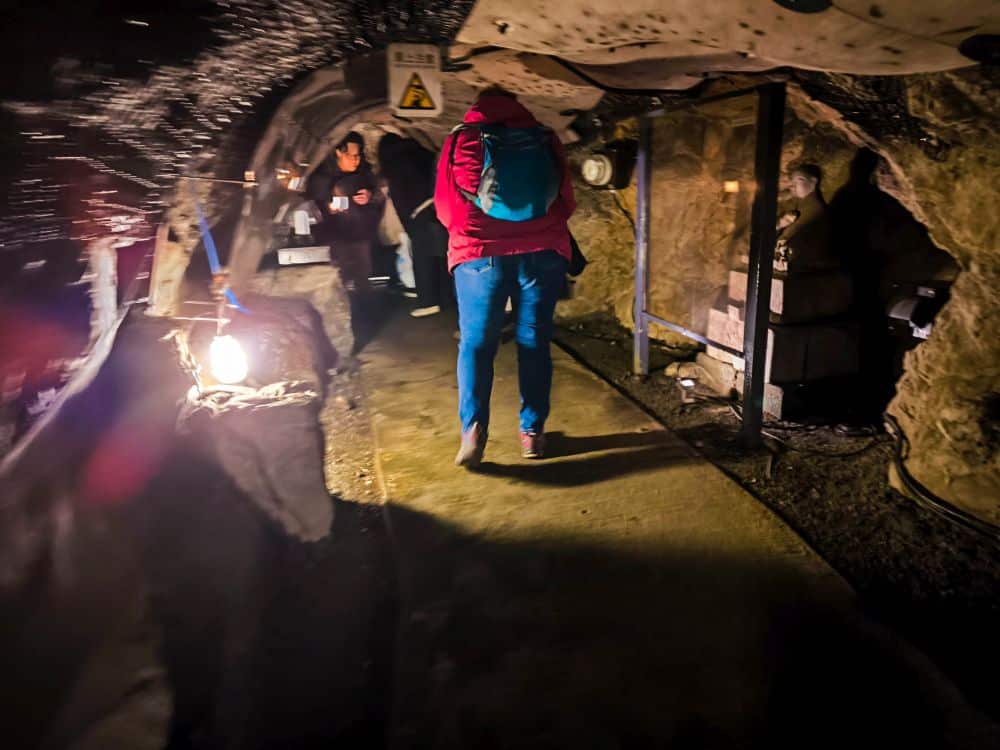 Eine Person mit einem blauen Rucksack geht durch den schwach beleuchteten Iwaya-Tunnel, dessen raue Felswände jeden Schritt widerhallen lassen. Lichter und Warnschilder säumen den Weg, andere folgen ihr.